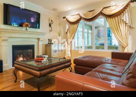 Rötlich-braunes L-förmiges Ledersofa und Couchtisch mit Glasplatte im Wohnzimmer mit Gaskamin im luxuriösen Haus. Stockfoto