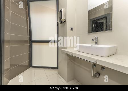 Kleines Badezimmer mit weißem Porzellanwaschbecken auf einer langen Arbeitsfläche aus hellem Stein und einem großen Fenster mit durchsichtigem Glas Stockfoto