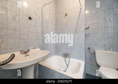 Altmodisches, gut abgenutztes Badezimmer mit blauen Fliesen, kleiner Badewanne und weißen Toiletten Stockfoto