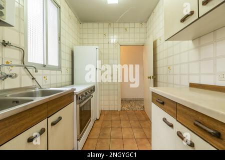 Alte Küche mit alten Möbeln in schlechtem Zustand, weißer Kühlschrank, alte Fliesen und Aluminiumfenster mit durchsichtigem Glas Stockfoto