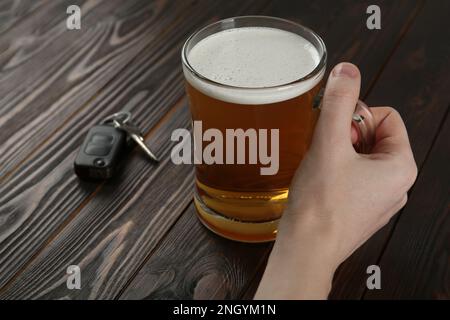 Ein Mann, der ein Glas alkoholisches Bier in der Nähe des Autoschlüssels am Holztisch hält, Nahaufnahme. Trunkenheit am Steuer ist gefährlich Stockfoto