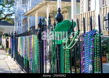 NEW ORLEANS, LA, USA - 13. FEBRUAR 2023: Selektivfokus des grünen Krewe of King Arthur hat auf einem eisernen Zaun mit Mardi Gras Perlen und Wohnhäusern in Uptown Stockfoto