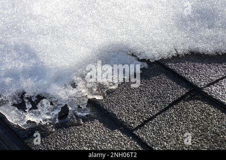Vollformat abstrakter Texturhintergrund von Schnee, der unter natürlichem Sonnenlicht auf einem Schindeldach aus Asphalt zu schmelzen beginnt Stockfoto