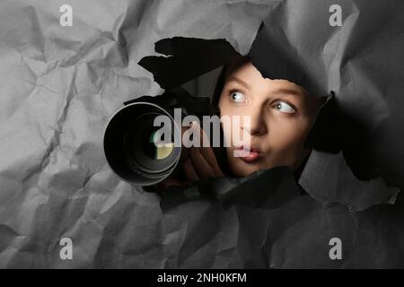 Versteckte Frau mit Kamera, die durch ein Loch in schwarzem Papier spioniert Stockfoto