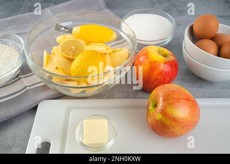 Backpfanne mit Apfelscheiben, Zitrone, Eiern, Zucker und Mehl in Schüsseln, Nahaufnahme auf einem Küchentisch. Apfelkuchenrezept, Zubereitungsprozess Stockfoto