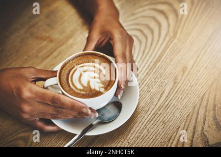 Du brauchst nur eine Umarmung im Becher. Aufnahmen aus dem hohen Winkel einer unbekannten Frau, die eine Tasse Kaffee hält, dekoriert mit Schaum. Stockfoto