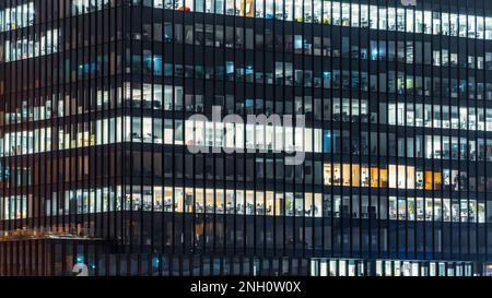 Nahaufnahme eines Bürogebäudes am Abend mit einigen beleuchteten Arbeitsplätzen. Moderner, verglaster Wolkenkratzer. Finanzdistrikt. Spät im Büro. Stockfoto