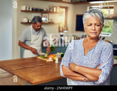 Er denkt, er sei ein besserer Koch als ich. Porträt einer erwachsenen Frau, die nach einem Streit mit ihrem Mann, der im Hintergrund kocht, aufgebracht wirkt. Stockfoto