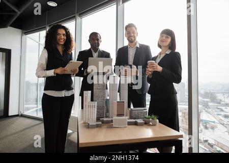 Ein Team aus vier verschiedenen, multiethnischen Geschäftsleuten, die sich im Sitzungssaal im Büro mit Panoramafenstern treffen, verschwommenes Stadtbild in der Nähe des Tisches mit einer Deckenverkleidungen aus Wolkenkratzern, die auf die Kamera schauen Stockfoto