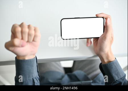Draufsicht eines Mannes, der ein weißes Smartphone-Mockup über dem weißen Schreibtisch hält und eine geballte Handbewegung zeigt. Sieg, Sieg, Erfolg, Angr Stockfoto