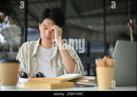 Ein unglücklicher und aufgebrachter junger asiatischer Student sitzt im Café oder in einem Co-Arbeitsplatz mit seinen Sachen und bereitet sich auf eine Prüfung vor. Stockfoto