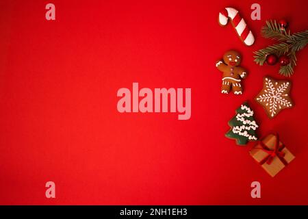 Süßer roter Hintergrund mit Keksen, Tannenzweig mit Weihnachtsbällen und ein Geschenk in handgemachtem Papier. Kekse in Form eines Lebkuchenmanns, Ch Stockfoto