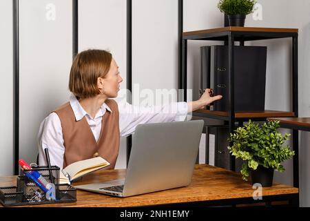 Eine Frau mittleren Alters nimmt Akten aus einem Regal in der Nähe ihres Schreibtisches Stockfoto