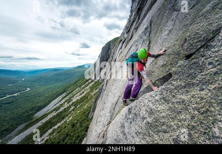 Ein Mann klettert steile Risse auf einer Klippe mit einem Tal dahinter Stockfoto