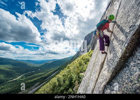 Ein Mann klettert steile Risse auf einer Klippe mit einem Tal dahinter Stockfoto