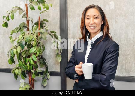Porträt einer positiven erwachsenen, ethnischen Unternehmerin im formellen Anzug, stehend mit einer Tasse heißes Getränk neben einem Topfbaum und schauend in die Kamera Stockfoto