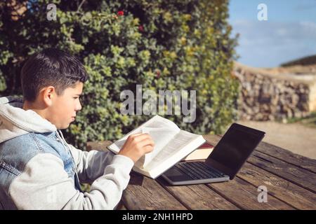 Junger Latinstudent blättert die Seite eines Buches vor einem Laptop um Stockfoto