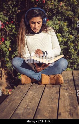 latina-Frau, die draußen sitzt, Musik hört und liest Stockfoto
