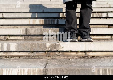 Fotografieren Sie Beine in Hosen und Turnschuhe auf Betontreppen im Freien. Personen- und Betontreppen, urbaner Stil. Stockfoto