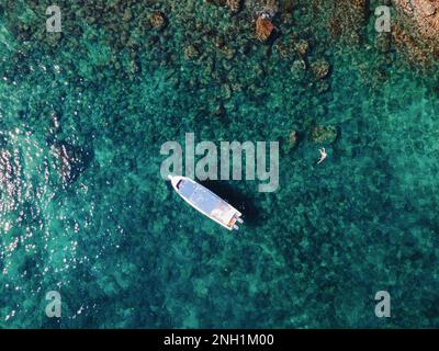 Ein weißes Boot, das auf einem ruhigen Gewässer vor einer felsigen Küste treibt, aus der Vogelperspektive Stockfoto