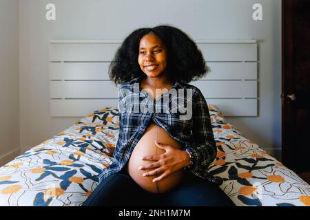 Eine junge Frau sitzt auf dem Bett und hält einen schwangeren Bauch Stockfoto