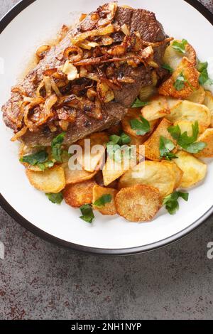 Traditionelles Zwiebelrostbraten deutsches Trockensteak mit Zwiebeln und Sauce, serviert mit gebratenen Kartoffeln in Nahaufnahme auf einem Teller auf dem Tisch. Vertikale Draufsicht Stockfoto