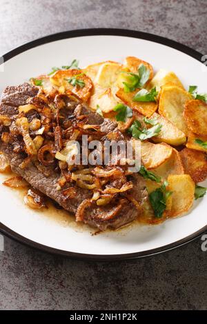 Traditionelles Zwiebelrostbraten deutsches Trockensteak mit Zwiebeln und Sauce, serviert mit gebratenen Kartoffeln in Nahaufnahme auf einem Teller auf dem Tisch. Vertikal Stockfoto