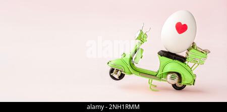 Kleine grüne Motorradfigur mit weißem Ei und kleinem roten Herz auf pastellrosa Hintergrund. Osterkonzept mit Kopierraum Stockfoto
