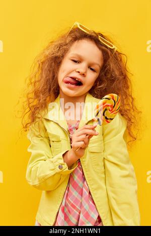 Positive Stimmung. Kleines süßes Mädchen, Kind mit lockigen Haaren in hellen Kleidern mit Lutscher auf gelbem Studiohintergrund. Der Begriff der Kindheit Stockfoto