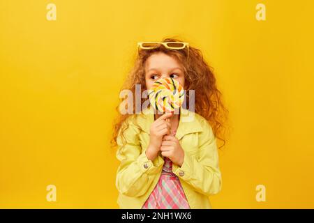 Versteckt sich hinter Lutscher. Kleines süßes Mädchen, Kind mit lockigen Haaren, das in hellen Kleidern auf gelbem Studiohintergrund posiert. Kindheitsbegriff, Emotionen Stockfoto