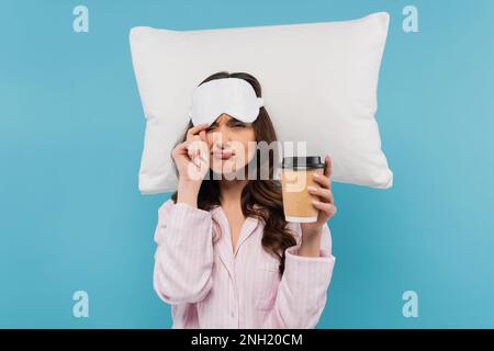 Unzufriedene Frau in Pyjamas und Schlafmaske hält Pappbecher mit Kaffee in der Nähe fliegendes Kissen isoliert auf blau, Stock Bild Stockfoto