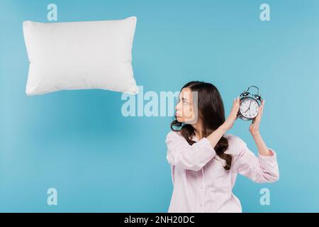 Verärgerte junge Frau in Pyjamas mit altem Wecker neben fliegenden Kissen auf blauem Stock Image Stockfoto