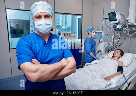 Porträt einer Krankenschwester mit gekreuzten Armen stehend auf der Intensivstation mit bewusstloser Patientin während der Pflege Stockfoto