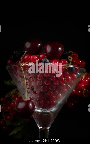 Auf einem dunklen Hintergrund in einem Glasbecher befinden sich rote Kirschen- und Johannisbeeren. Stockfoto