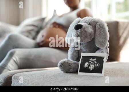 Bald kommt ein süßes kleines Wunder. Nahaufnahme eines Sonogramms neben einem ausgestopften Teddybär mit einer schwangeren Frau im Hintergrund. Stockfoto