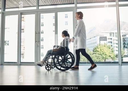 Ein anderer Patient wurde erfolgreich behandelt. Eine junge Ärztin schubst einen Patienten im Rollstuhl in einem Krankenhaus. Stockfoto