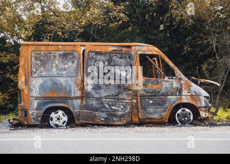 Der große Van neben der Straße ist ausgebrannt. Das Auto nach dem Unfall. Ein ausgebranntes Auto ohne Fenster und Reifen Stockfoto