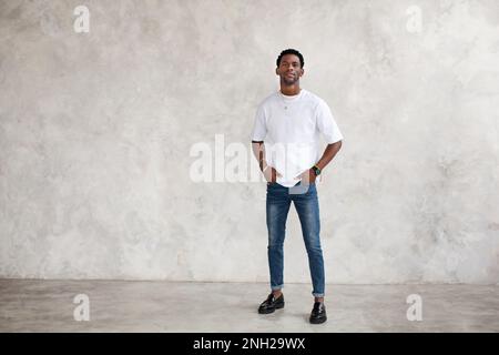 Ein afroamerikanischer junger Mann steht in voller Länge vor einer hellen texturierten Wand im Studio. Das männliche Model hat die Hände in den Taschen und trägt lässige Kleidung in Weiß Stockfoto