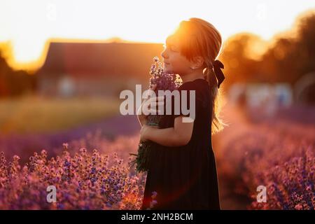 Porträt eines niedlichen Mädchen riecht ein Lavendel Blumen. Ein Kind läuft bei Sonnenuntergang in einem Lavendelfeld. Kind in schwarzem Kleid hat Spaß auf Natur Stockfoto