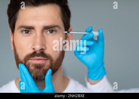 Ein Mann mittleren Alters erhält ein Schönheitsfoto von einem Kosmetiker mit grauem Studiohintergrund, freier Platz Stockfoto