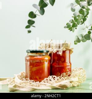 Probiotische Lebensmittel. Umweltfreundliches Stillleben mit eingelegtem oder fermentiertem Gemüse. Lecho und Ajvar in Glasgefäßen auf Netzbeutel auf blauem Hintergrund mit eucal Stockfoto