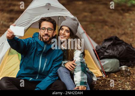 Fröhlicher weißer Mann und Frau der Jahrtausendwende ruht sich im kalten Wald aus, genießt das Abenteuer, sitzt im Zelt, macht Selfies Stockfoto
