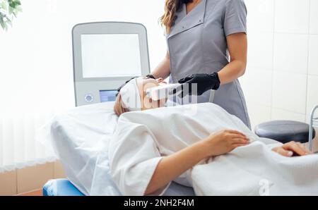 Porträt einer entspannten jungen Klientin, die eine SMAS Ultraschall-Gesichtsliftmassage mit professioneller Ausstattung im Beauty Center erhält Stockfoto