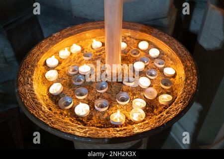 Religiöse Teelichter auf einem Sandfeld in einer englischen Kathedrale während eines Gottesdienstes. Eine große Kerze in der Mitte ist teilweise zu sehen. Stockfoto