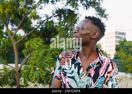 Junger afrikanischer Mann mit kurzen Haaren und Brille, sitzt auf einem Platz und beobachtet die Umgebung um ihn herum, kopiert Raum. Stockfoto