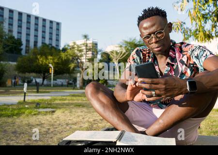 Nutzt mobile Apps, um Ihren Tag zu organisieren, ein junger Mann afrikanischer ethnischer Herkunft mit Brille, farbenfrohem Hemd und Shorts sitzt draußen mit seinem Handy, Kopie Stockfoto