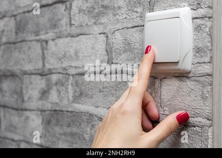 Weibliche Hand drückt die Finger am Lichtschalter. Die Frau macht das Licht im Raum an. Der Schalter wird mit dem Finger gedrückt. Stockfoto