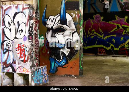 Graffiti mit weißer Gesichtsmaske, blauen Hörnern und roten Augen und andere Graffiti bedecken die Wände in einem alten verlassenen Gebäude. Stockfoto