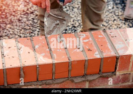 Nahaufnahme einer Mauer aus Ziegelstein und einer Kelle, die der Arbeiter zum Auftragen und Glätten des Mörtels zwischen Ziegelsteinen verwendet. Maurer, der den letzten Schliff auf den Ziegelstein legt Stockfoto