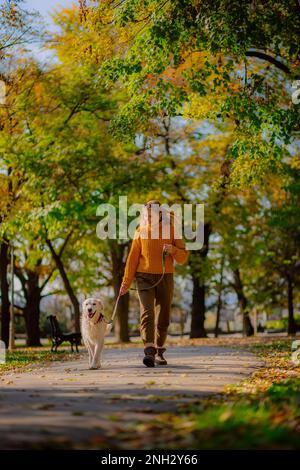 Belgrad, Serbien. 10. November 2022. Fröhliche junge Frau, die an einem sonnigen Tag mit ihrem goldenen Retriever in einem Park spaziert. Stockfoto